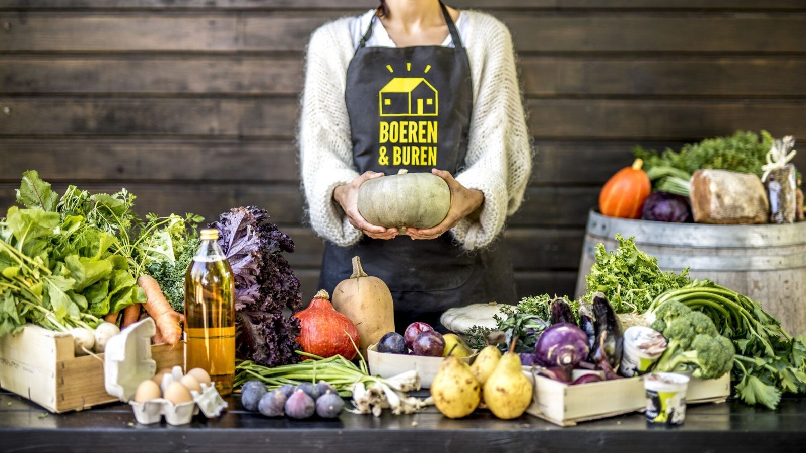 Persoon in wit vest en zwart schort met geel logo houdt pompoen vast, omgeven door groenten, fruit en olie op tafel