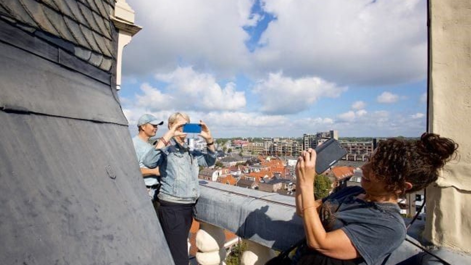 Twee personen op een dak nemen foto's van een stadsgezicht met rode daken en flatgebouwen onder bewolkte hemel