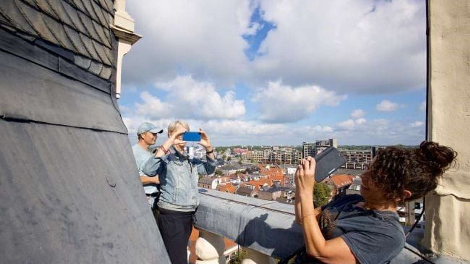 Twee personen op een dak nemen foto's van een stadsgezicht met huizen en wolkenlucht, terwijl een derde persoon hen fotografeert.
