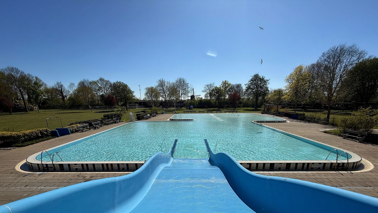 Rechthoekig buitenbad met turquoise water en twee blauwe glijbanen, omgeven door houten terras, grasvelden en loofbomen op zonnige dag.