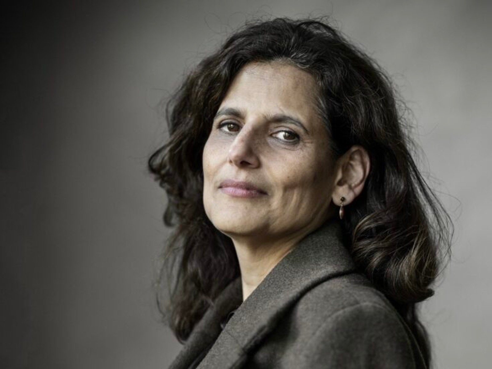 Portretfoto vrouw met donker golvend haar en grijze blouse tegen grijze achtergrond, kijkend naar camera