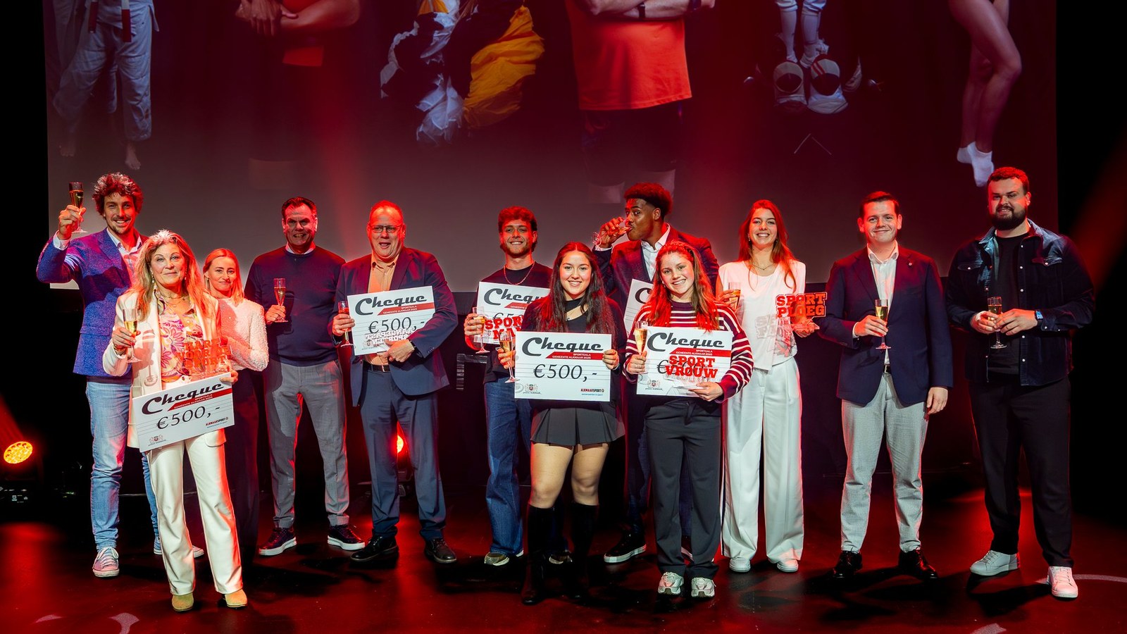 Groep personen op podium onder rood licht, enkele houden cheques van 500 euro omhoog