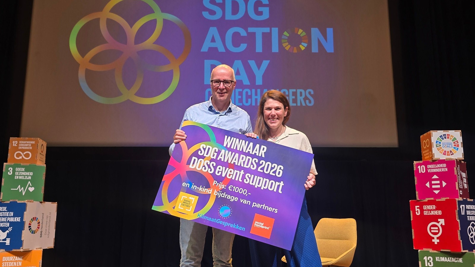 Twee personen houden SDG Awards 2026 DOSS Event Support banner vast op podium met kleurrijke SDG-logo's