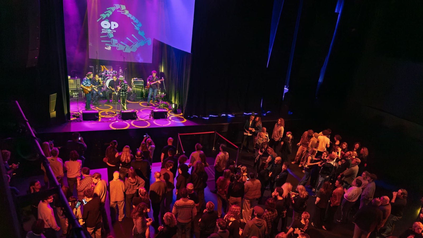 Podiumoptreden met band en publiek in paarse en blauwe verlichting in theater