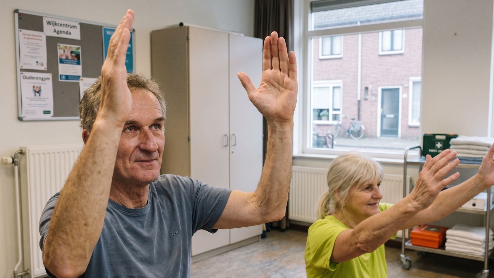 Man en vrouw voeren armbeweging uit in een gymnastiekzaal met ramen, agenda's op een prikbord zichtbaar
