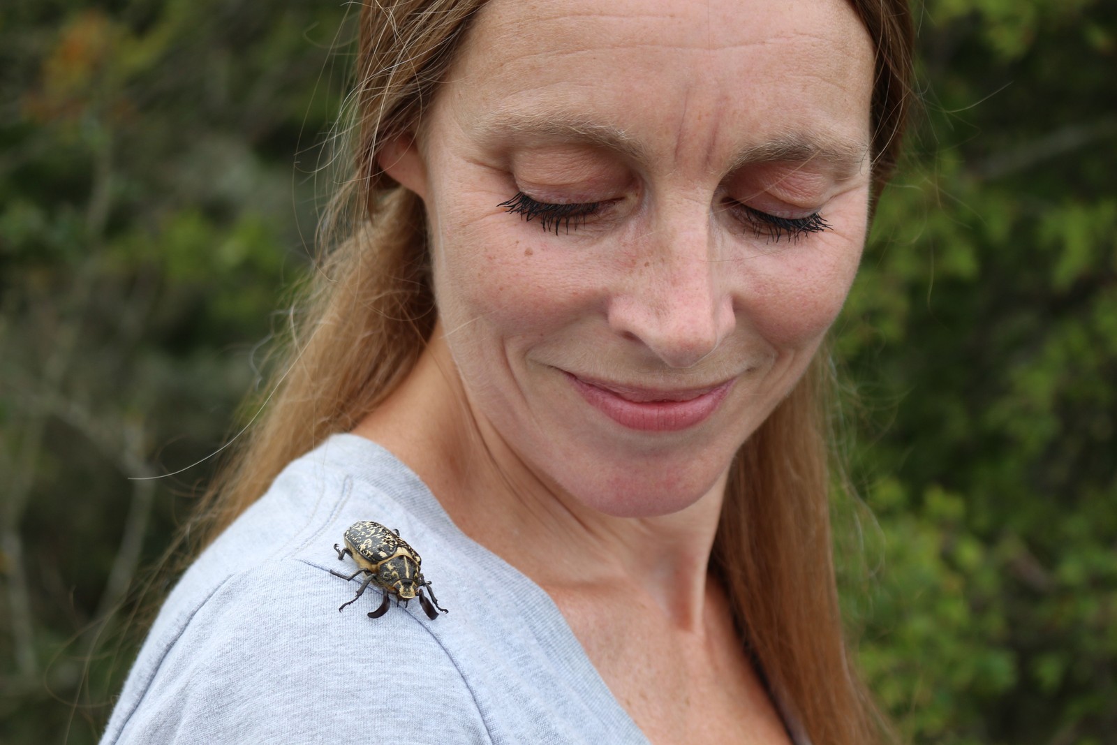 Vrouw met rood haar en gesloten ogen draagt een lichtgrijs shirt met een insect erop. Op de achtergrond groen gebladerte.