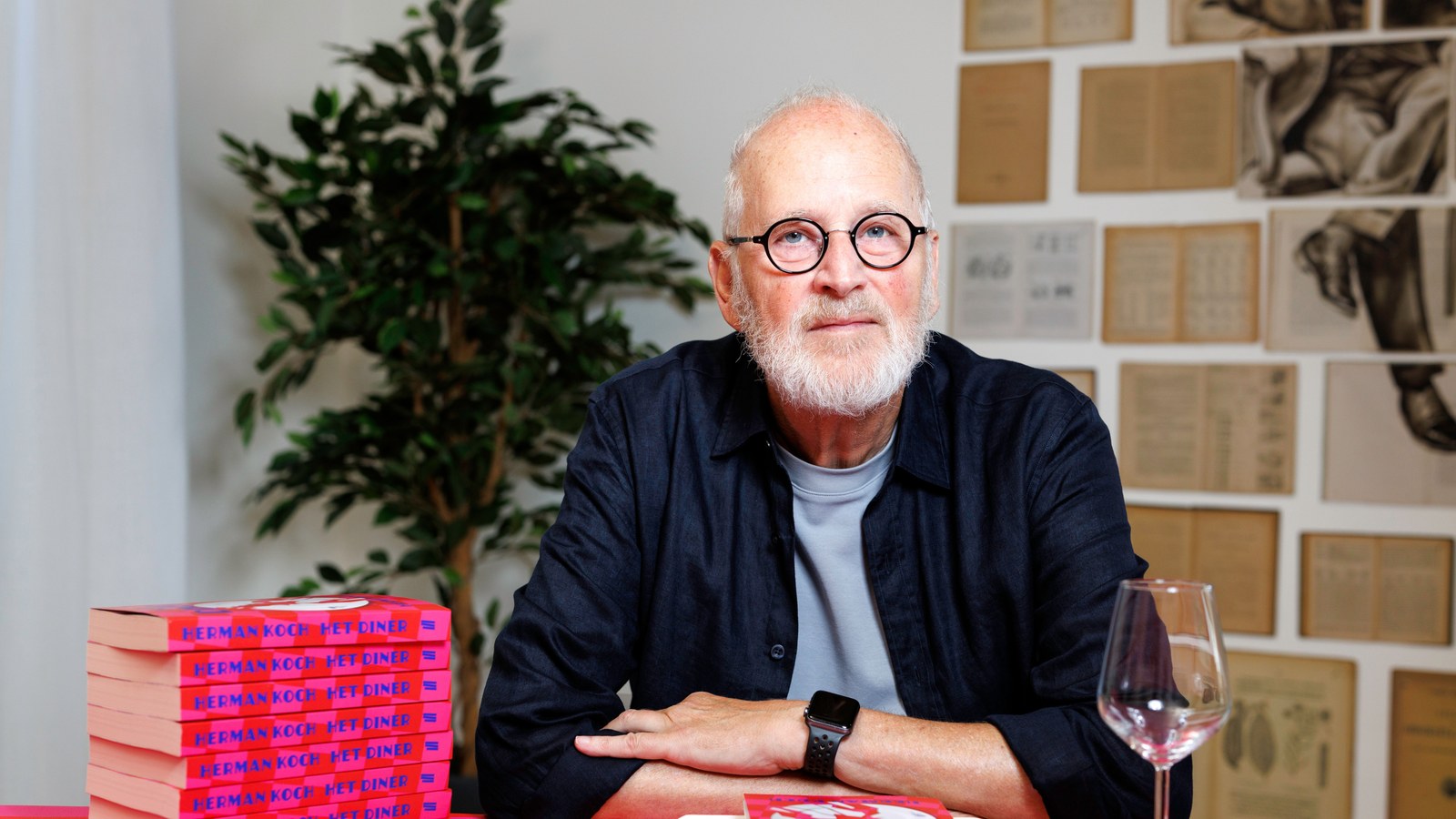 Man met bril en wit baard aan rode tafel met stapel rode boeken, wijnglas en bord. Achtergrond met plant en boekenrek.