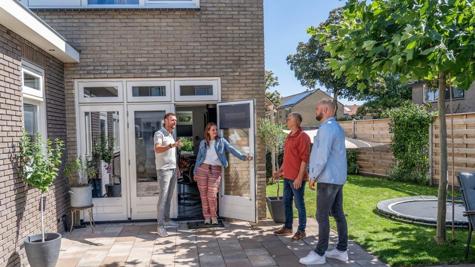 Vier personen converseren buiten baksteenhuis bij open tuindeuren met terras en trampoline zichtbaar