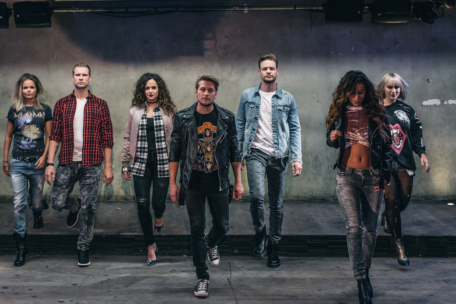 Zeven personen in casual en rockstijl kleding lopen naar de camera toe tegen een grijze betonnen muur met spotlights erboven.