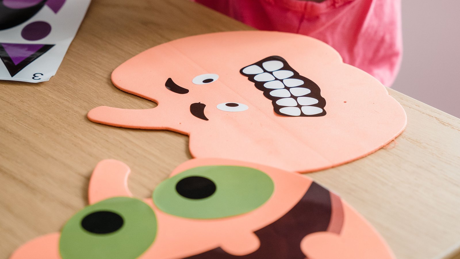Kind in roze shirt bestudeert twee gekleurde kartonnen gezichten met grote ogen op houten tafel. Een gezicht is perzikkleurig met witte tand