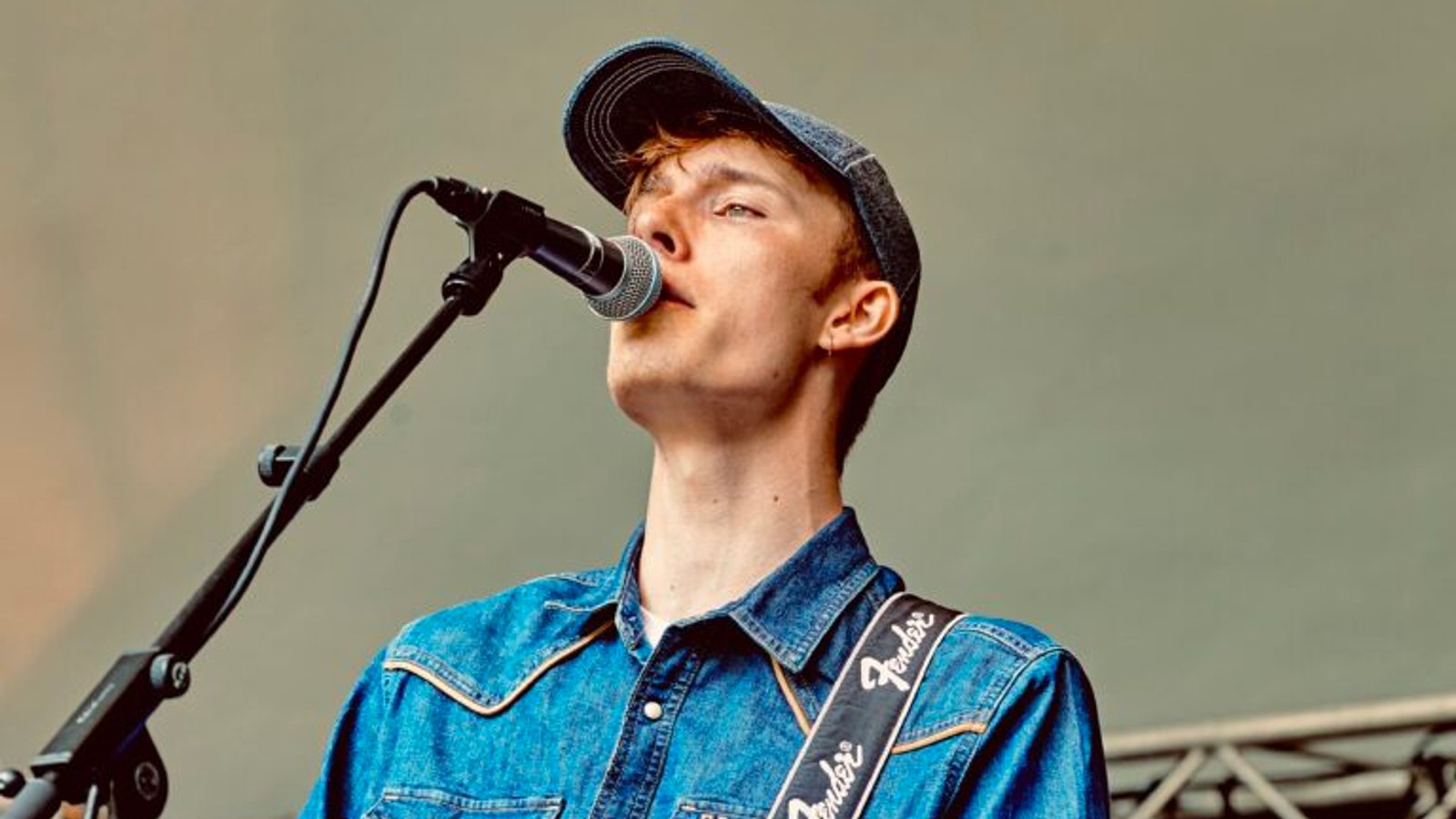Muzikant zingt in microfoon en bespeelt elektrische gitaar, draagt blauw denim overhemd en pet.