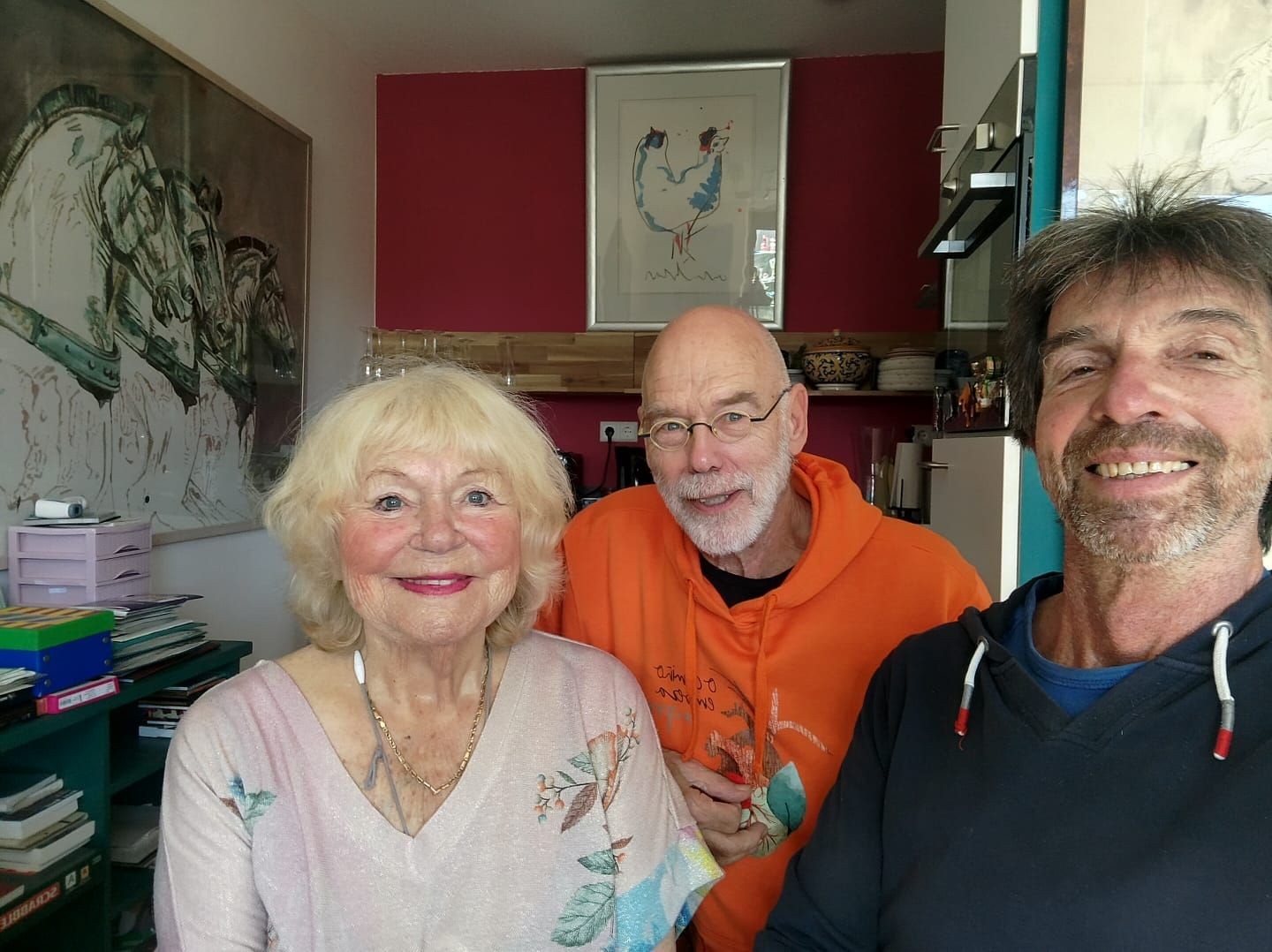 Drie personen poseren in een kunstenaarsstudio met rode en witte muren, kunstwerken aan de wand en kunstboeken op shelves.