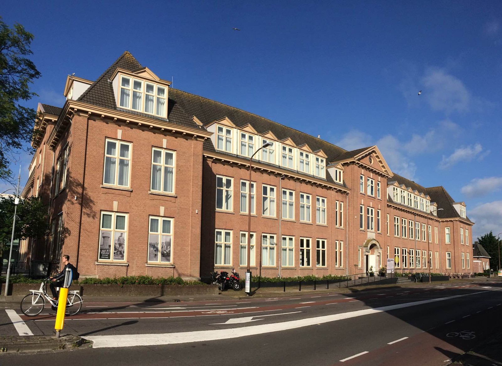 Rood bakstenen schoolgebouw met mansardedak en witte raamkozijnen aan een straat met fietspad. Een witte fiets staat vastgemaakt aan een gel