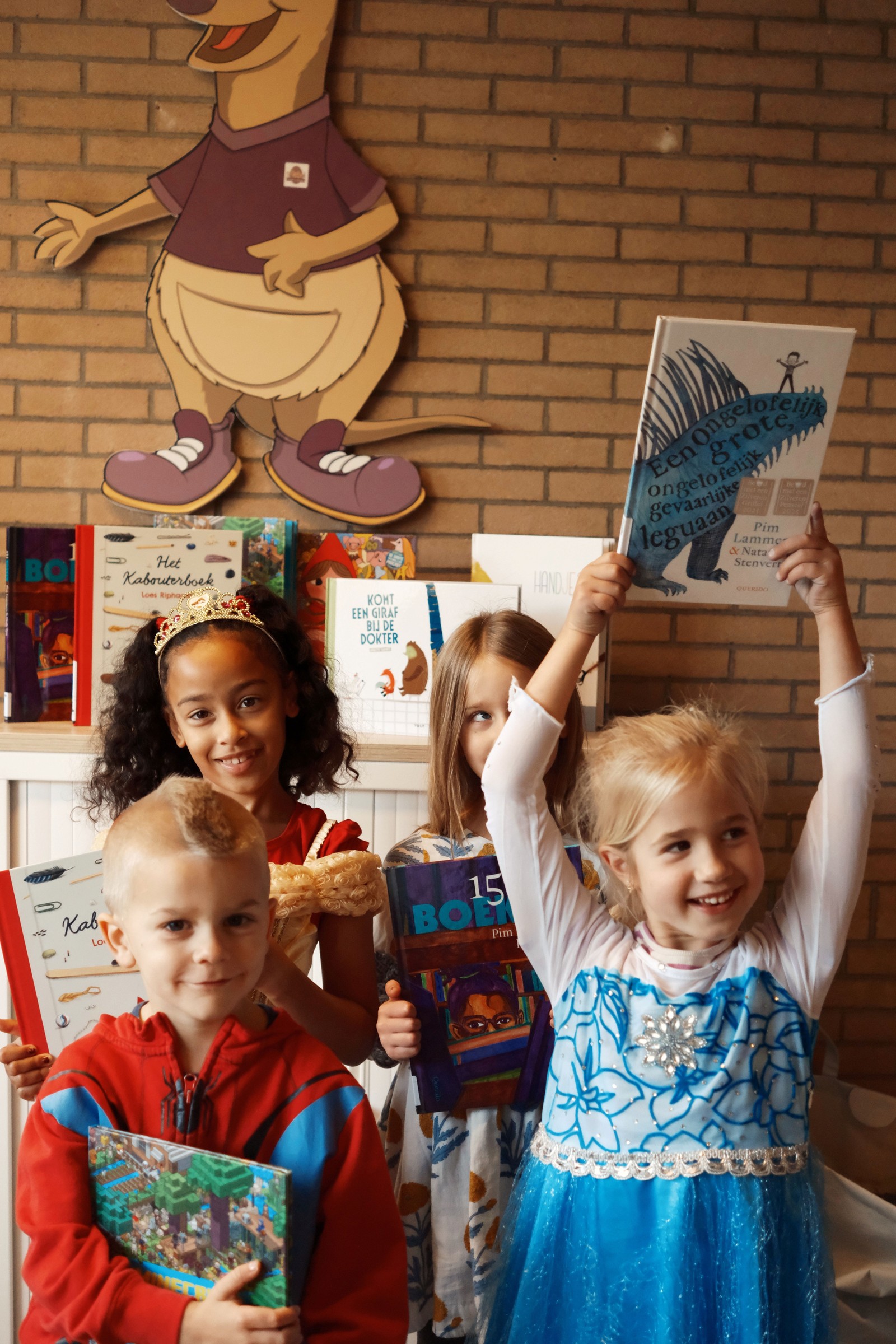 Vier kinderen houden kinderboeken vast voor een bakstenen muur met een groot cartoon-karakter erop. Ze dragen feestelijke kleding en één kin