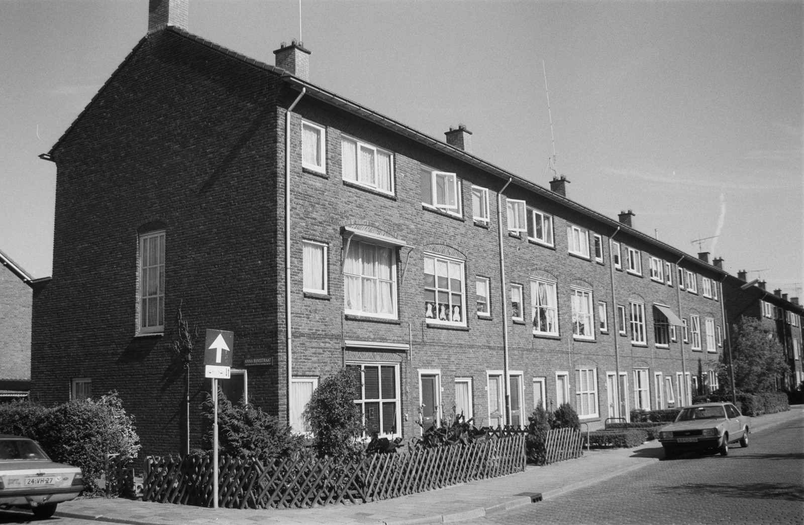 Rij van bakstenen woningen met drie verdiepingen aan een straat. Voor de huizen staan auto's geparkeerd en lage houten hekken. Zwart-witfoto