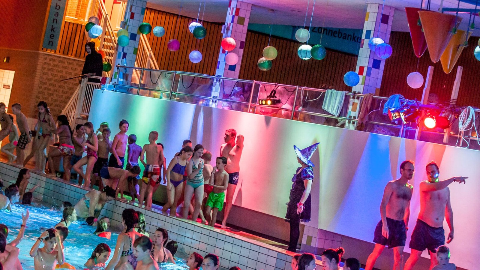 Veel kinderen zwemmen in een binnenbad met gekleurde verlichting en dansende figuren op een projectiescherm, kleurrijke ballonnen hangen