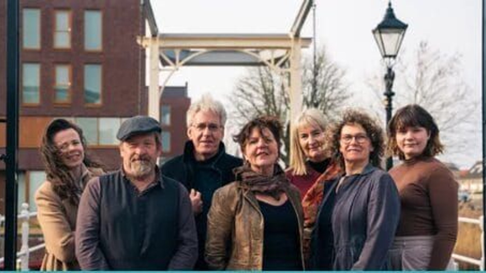 Zeven personen poseren buiten voor een houten constructie en straatlantaarn met gebouwen op achtergrond