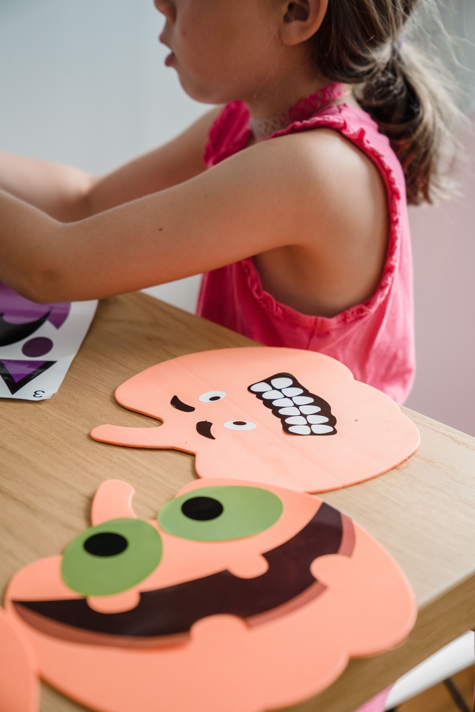 Kind in roze shirt werkt aan gekleurde kartonnen gezichtsfiguren op houten tafel. Onderaan oranje gezicht met groene ogen en glimlach, erbov