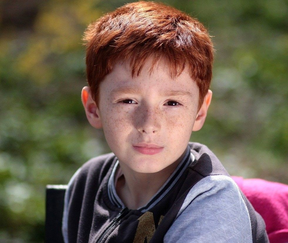 Jongen met rood haar en sproeten, draagt een donkerblauw en grijs jack, tegen een vervaagde groene achtergrond.