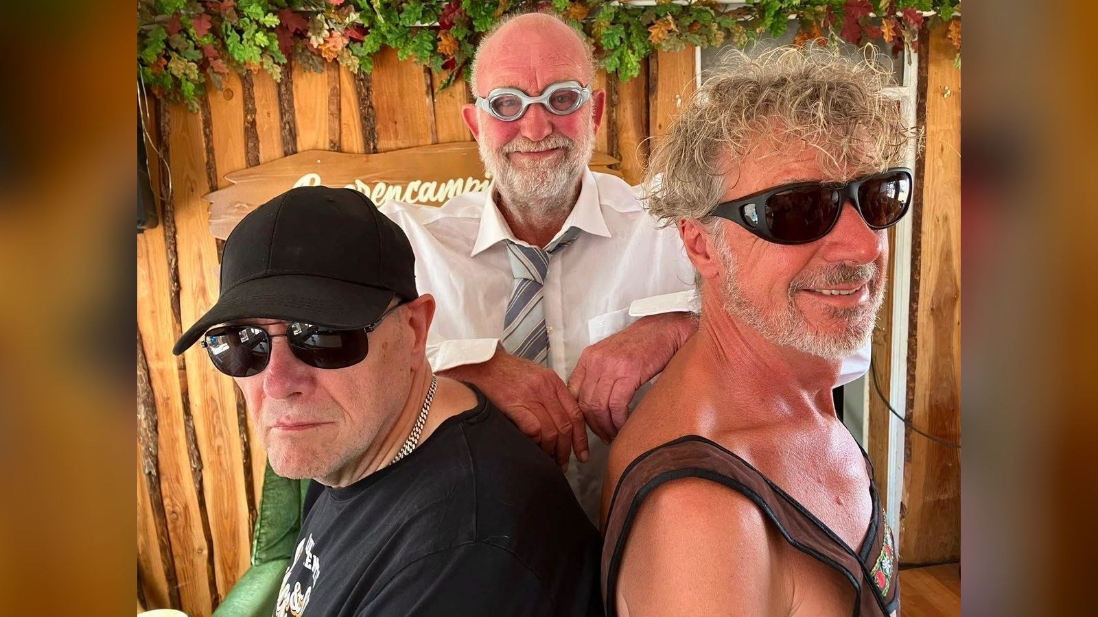 Drie mannen poseren samen in een houten gebouw met plantendecoratie. De man links draagt een zwart petje en zonnebril, de man in het midden