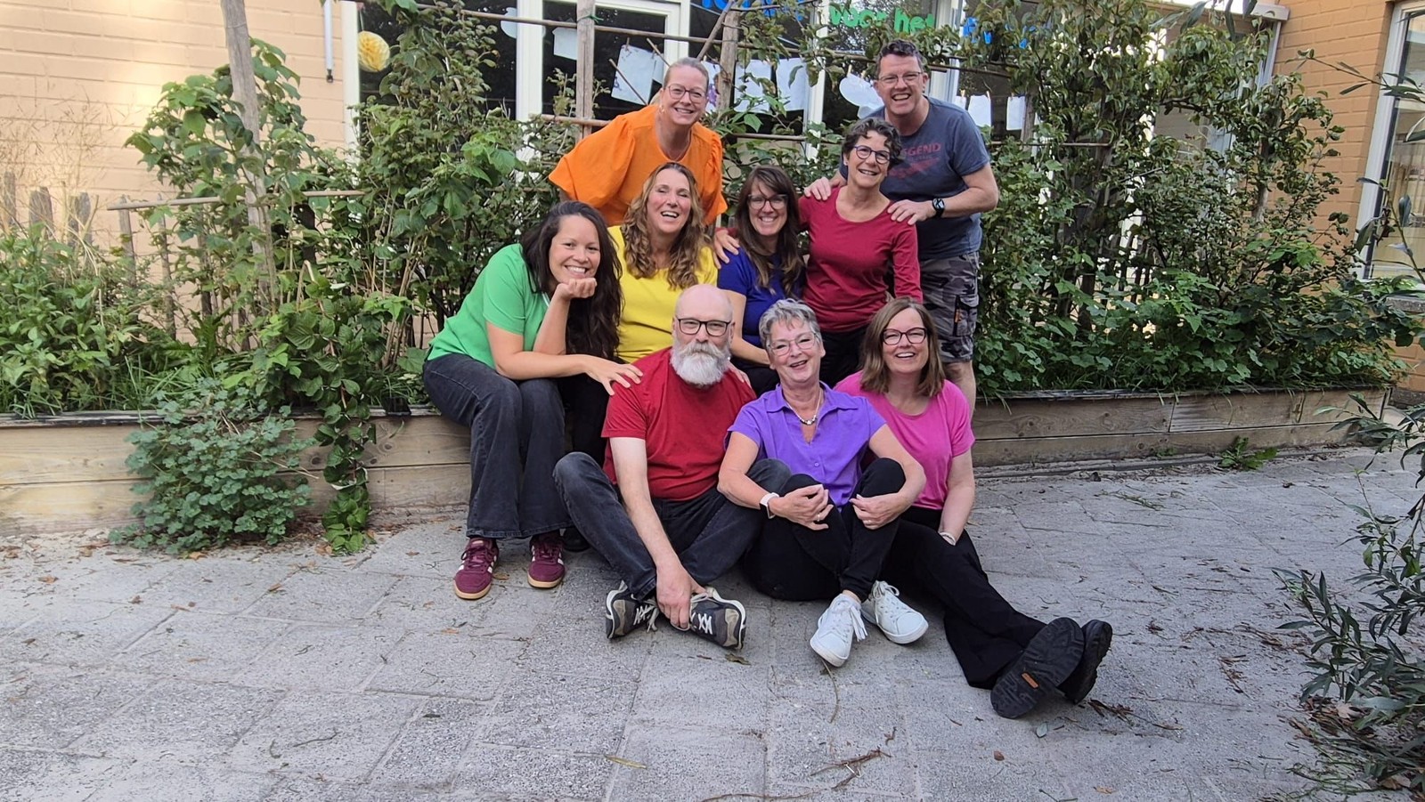 Groep van negen personen poseren samen in een tuin met groene planten en klimop tegen een bakstenen muur.