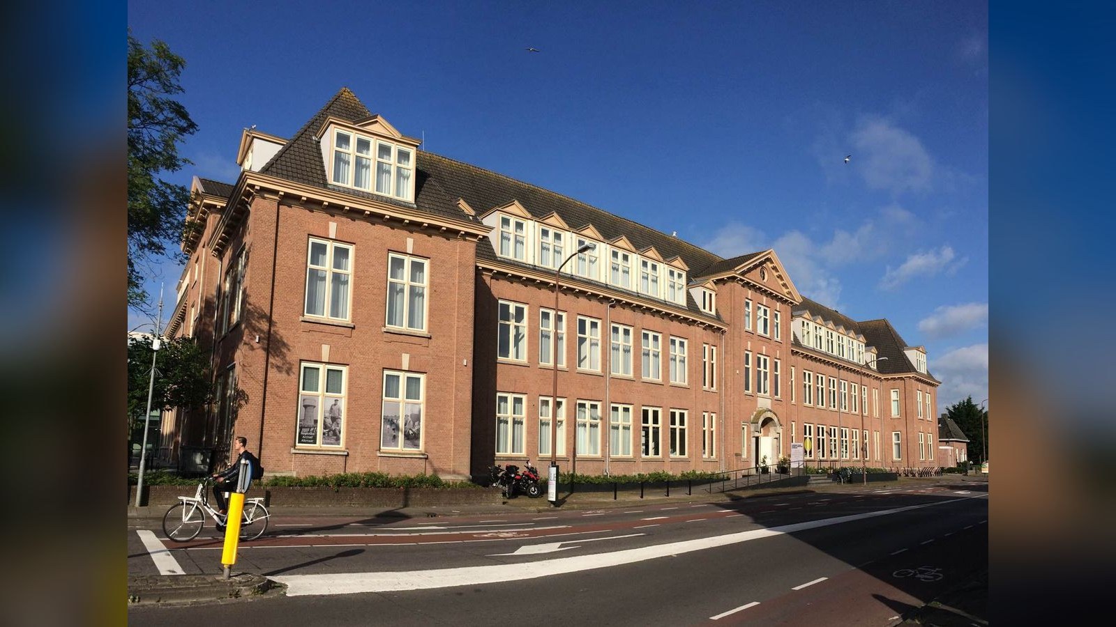 Rood bakstenen gebouw met mansardedak en witte raamkozijnen. Voor het gebouw fietser met gele fietsslot, straat met fietspaden en parkeerpla