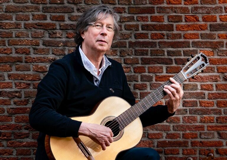 Man met bril speelt klassieke gitaar tegen bakstenen muur, draagt donker vest over gestreept overhemd.