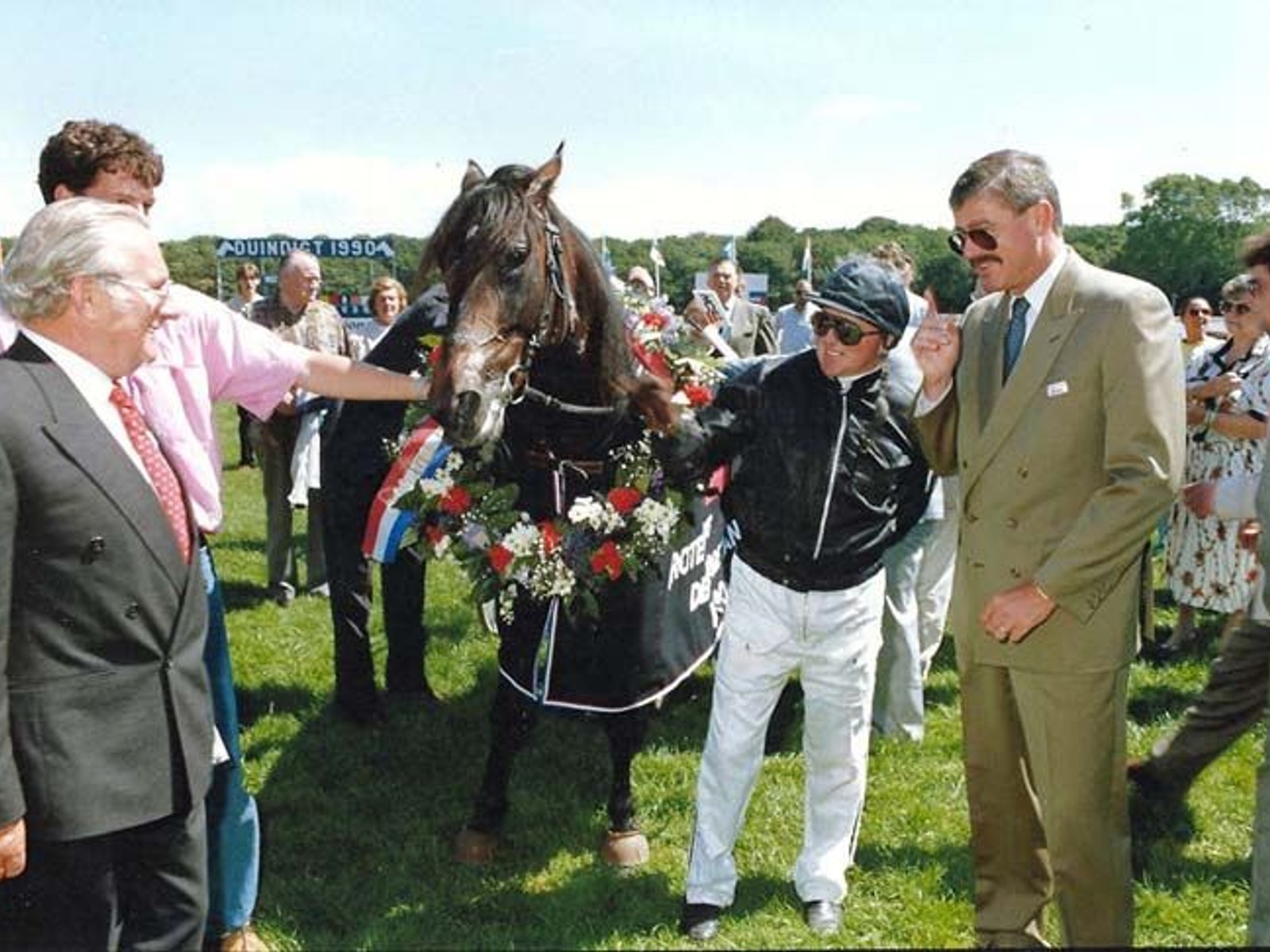 Zwart paard met bloemversiering staat op grasveld omringd door mannen in pak en jockey in ruiterskleding bij officiële paardenraceceremonie.